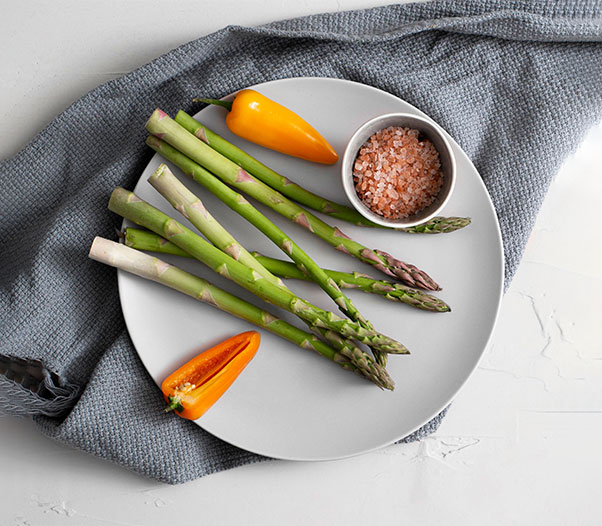 Fresh green asparagus spears arranged on a white plate with colorful peppers and a small bowl of Himalayan salt bulk, styled on a gray cloth for a clean, natural food presentation.